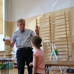 Mężczyzna trzyma w ręce słoik z kukurydzą, z którego wyciągnął piłeczkę. Pokazuje ją stojącej przed nim dziewczynce