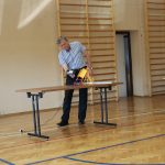 Mężczyzna przy stoliku trzyma w ręce suszarkę, której powietrzem napędza stojący na stole pojazd z klocków