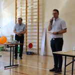 W sali stoi dwóch mężczyzn, jeden mówi do mikrofonu, drugi się przygląda