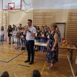 Mężczyzna stoi na sali gimnastycznej, obok niego kobieta. Za nimi grupa uczniów