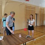 Uśmiechnięta dziewczynka stoi przy stoliku, nad którym pochyla się nauczyciel