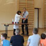 Dziewczynka w okularach ochronnych i białym fartuchu nalewa do naczynia wodę. Obok stoi nauczyciel