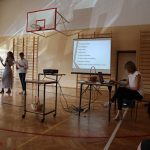 Na ekranie wyświetlone słowa piosenki, przy stoliku siedzi kobieta, w oddali stoi chłopiec i dziewczyna